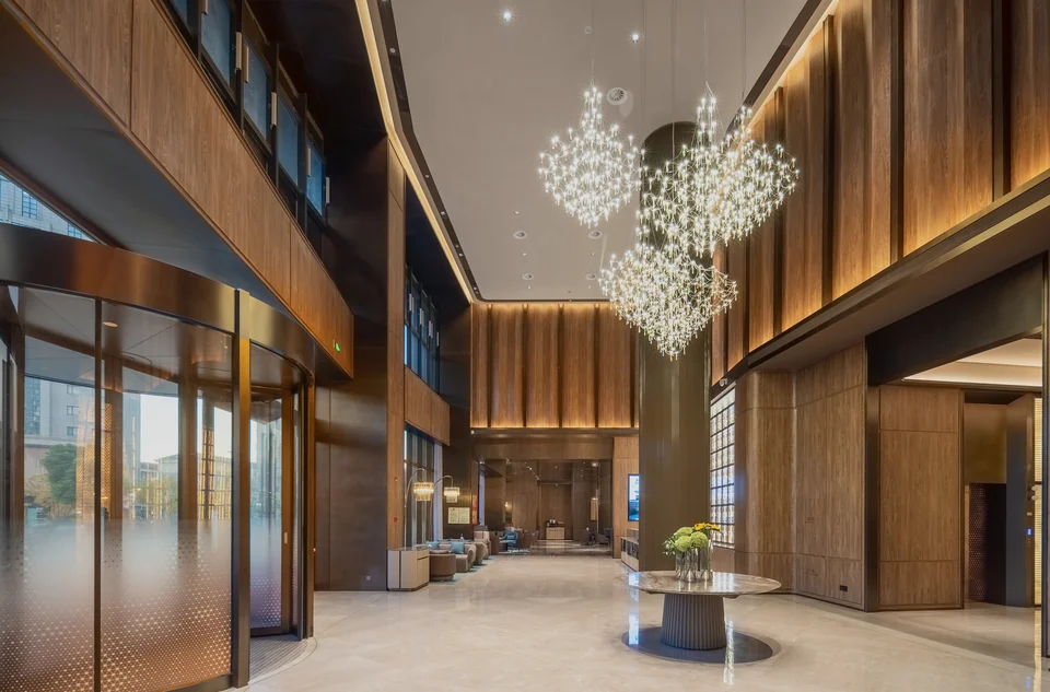 Completed double-height hotel lobby with warm-toned wall panels, pendant lights and a revolving entrance in Shaoxing.