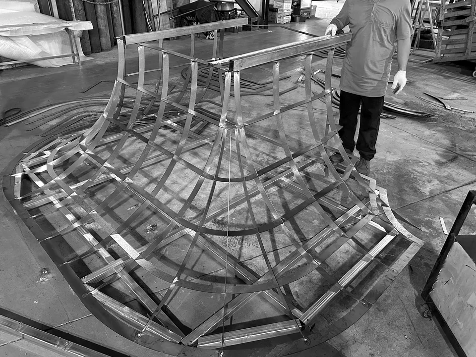 Workshop fabrication view of an open curved frame with ribs, joints and a worker for scale.