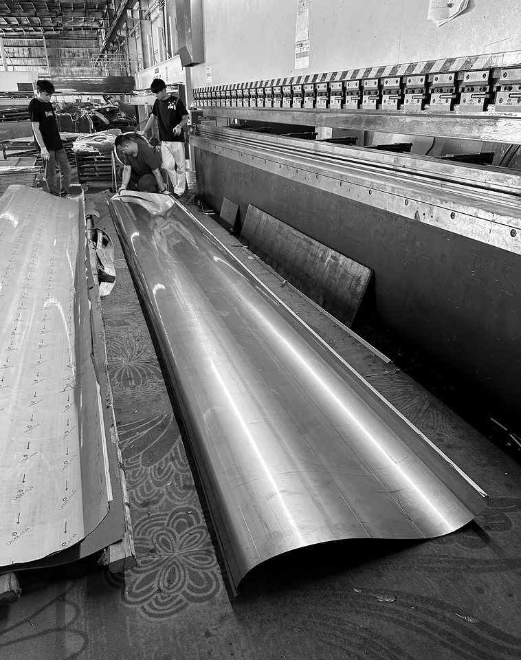 Workshop fabrication view of a long curved metal sheet beside bending equipment and workers.