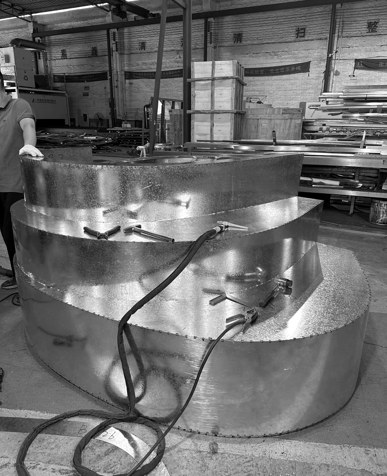 Workshop fabrication view of a layered curved metal component with weld points, tools and a worker.