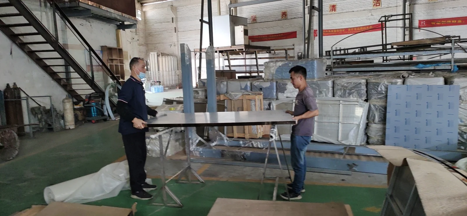 Workers handling a large metal-faced panel at a workshop table.
